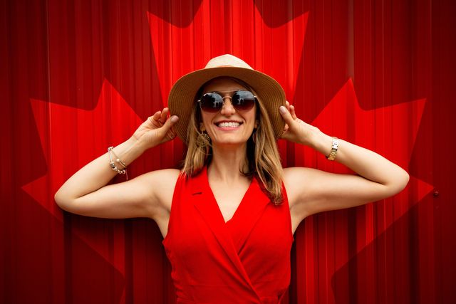 Favoritos para listas de regalos de Canadá, woman dressed in a red dress, sunglasses, and a hat standing in front of a red curtain with a bright red maple leaf on it.